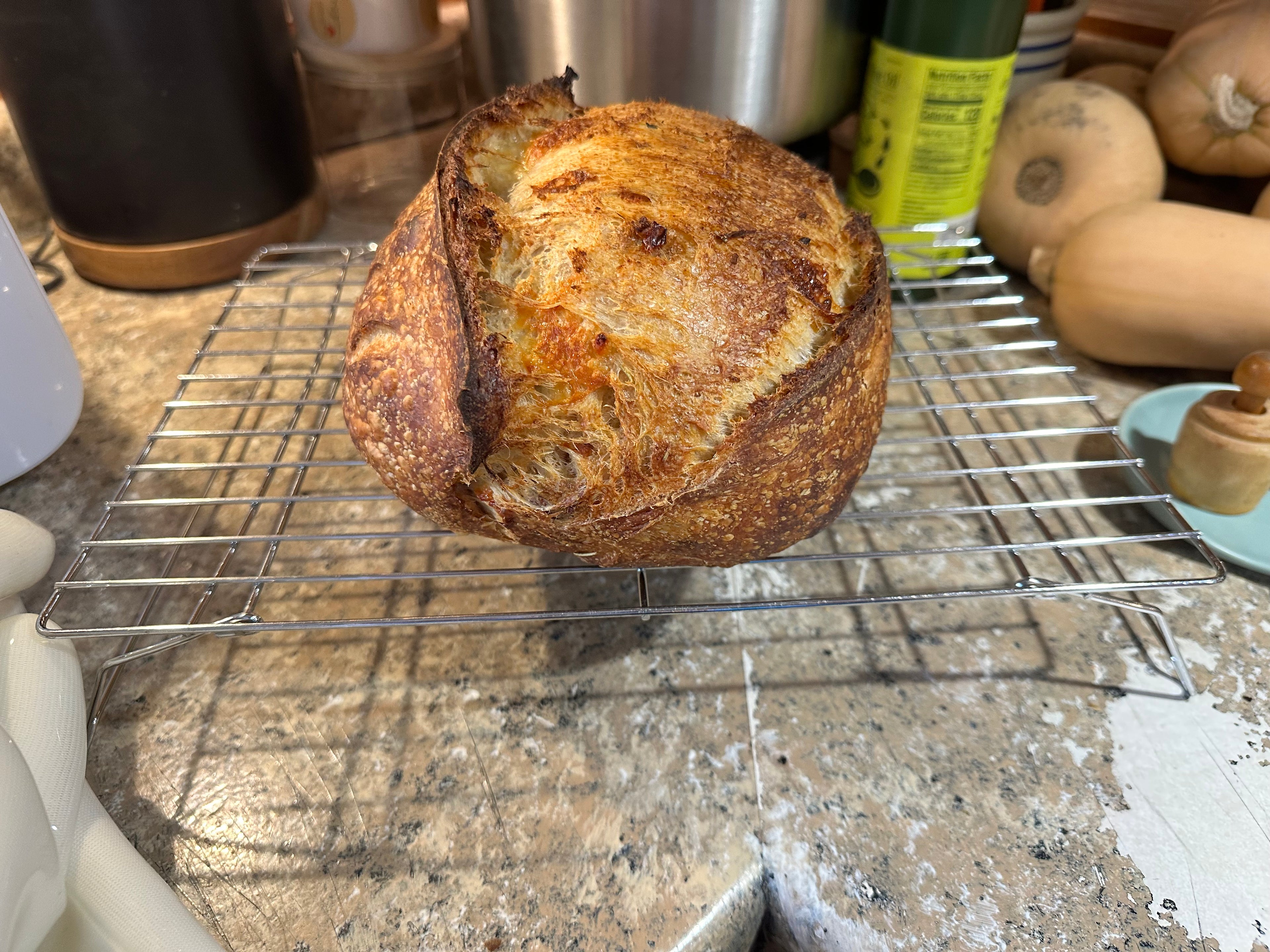 A freshly baked sourdough bread loaf with a golden-brown crust sits on a metal cooling rack on a kitchen countertop.