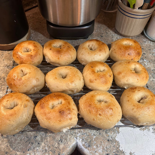 Twelve golden-brown baked bagels arranged on a wire cooling rack on a speckled granite kitchen countertop, with kitchen appliances and containers visible in the background.